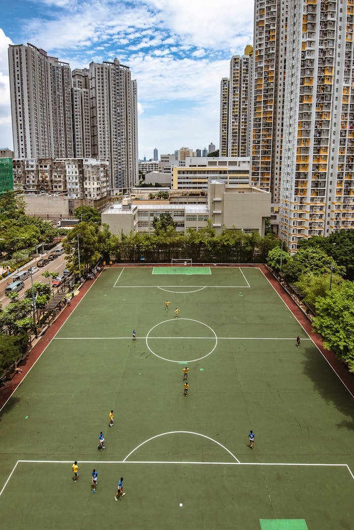 Aerial view of a football game on an urban field, surrounded by tall buildings and blue sky.