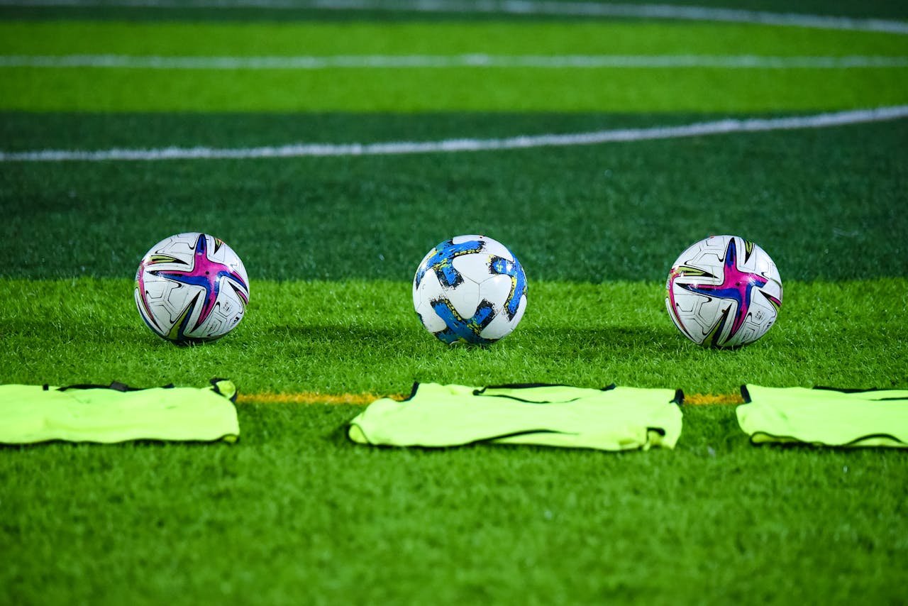 Three soccer balls and training bibs on a lush green field in Rawalpindi, Pakistan.